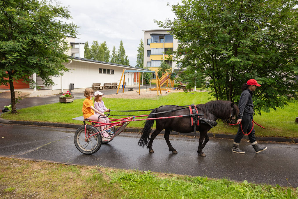 Kuvassa poni vetää kärryjä, joissa istuu kaksi lasta. Ponia taluttaa henkilö punaisella narulla, ja taustalla näkyy asuinalueen rakennuksia ja leikkikenttä. Tapahtuma vaikuttaa olevan rauhallinen ja ulkoilmassa tapahtuva aktiviteetti, jossa lapset pääsevät kärryajelulle ponin kanssa.