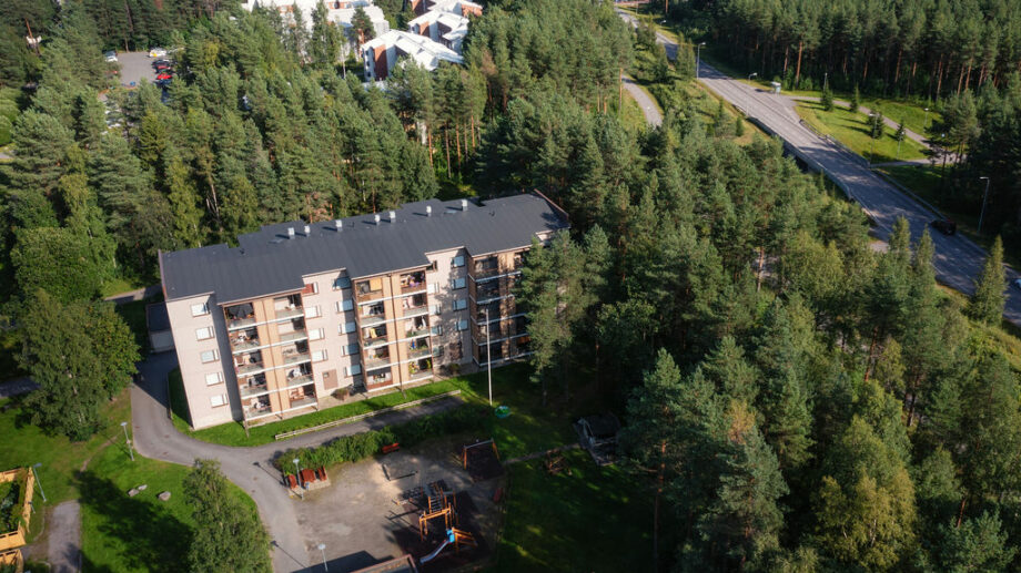 Ilmakuva asuinrakennuskompleksista, joka sijaitsee metsän keskellä. Kerrostaloa ympäröivät puut, ja pihalla on pieni leikkipaikka. Talo sijaitsee lähellä isompaa tietä.