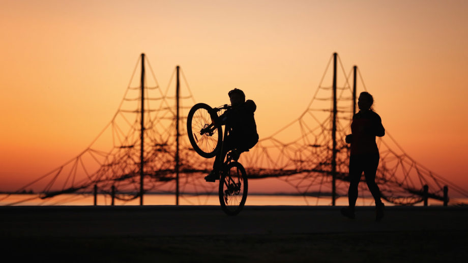 Poika temppuilee pyörällä, äiti lenkkeilee vieressä. Taustalla upea auringonlasku.