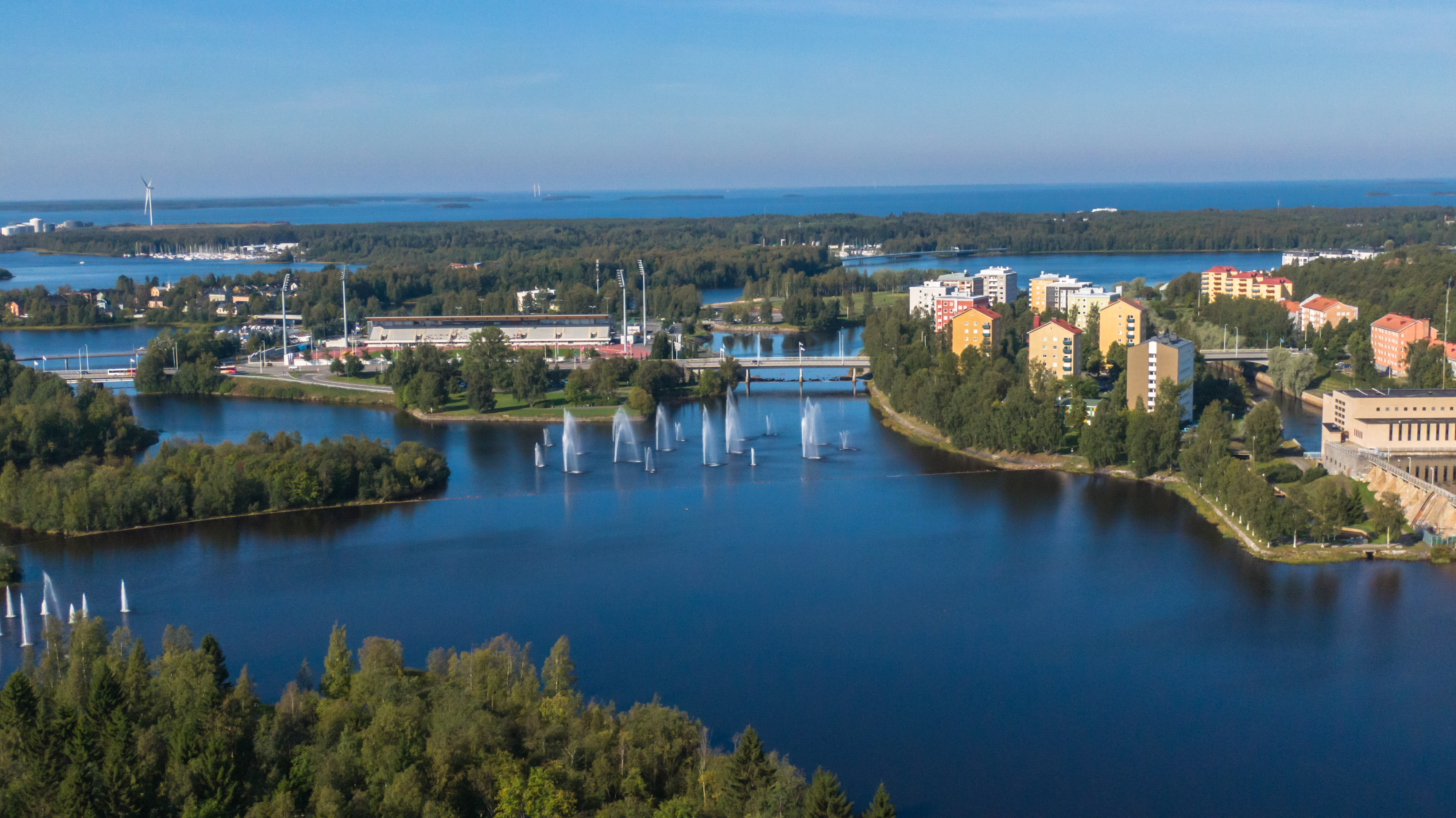 Maisemakuva lintuperspektiivistä, jossa on Oulujoen suistoa ja Toivoniemen asuinalue. Kerrostaloja, sininen taivas ja kesäistä kaupunkia.