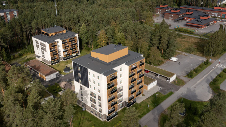 Ilmakuva kuusikerroksisesta kerrostalosta, ympärillä puita ja muita rakennuksia.