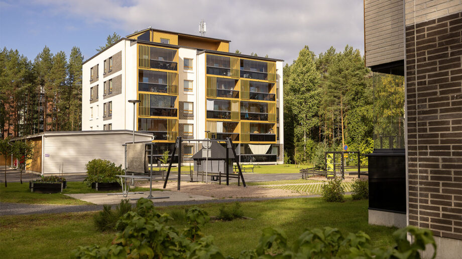 Leikkipiha ja kerrostalo metsäisessä ympäristössä, hiekkakenttä ja keinu etualalla.