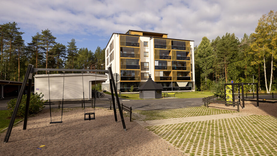 Leikkipiha ja kerrostalo metsäisessä ympäristössä, hiekkakenttä ja keinu etualalla.