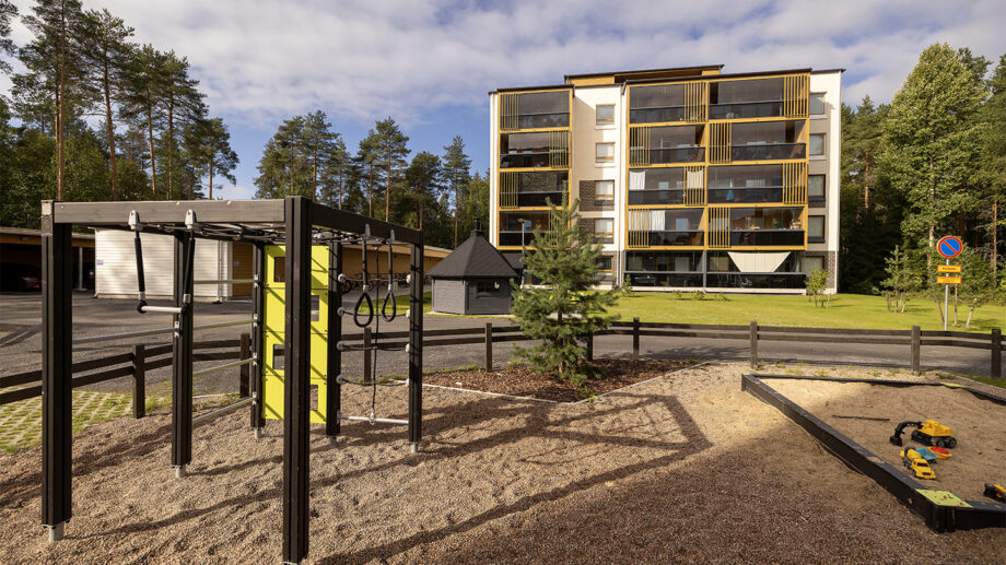 Hiekkapohjainen leikkipiha, keinuja ja liukumäki, taustalla moderni kerrostalo.
