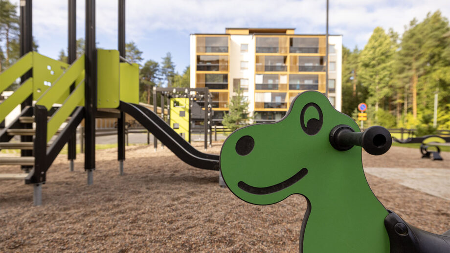Leikkipiha, vihreä dinosauruslaite ja taustalla kuusikerroksinen asuinkerrostalo.