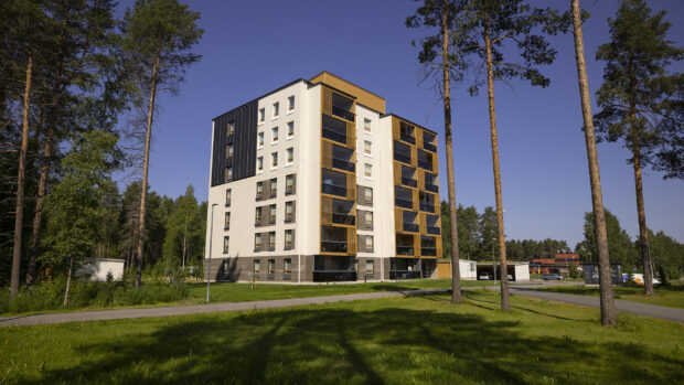 Kuvassa on moderni kerrostalo vehreässä ympäristössä. Rakennus on kuusikerroksinen ja sen julkisivussa on vaaleita ja tummia elementtejä, mikä luo tyylikkään ja nykyaikaisen ilmeen. Talon ympärillä on korkeat puut ja hyvin hoidettu nurmikko, ja taivas on kirkkaan sininen, mikä antaa kuvalle aurinkoisen ja rauhallisen tunnelman.