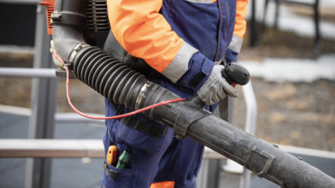 Kuvassa on henkilö, joka pitää lehtipuhallinta käsissään. Hänellä on päällään kirkkaan oranssi ja sininen työhaalari sekä hanskat. Kuvan keskipiste on lehtipuhaltimessa ja henkilön työvarusteissa. Tausta on hieman epätarkka, mutta viittaa ulkotyöympäristöön, mahdollisesti puisto- tai katualueella. Tunnelma on toiminnallinen, ja kuva korostaa työntekoa ulkona.