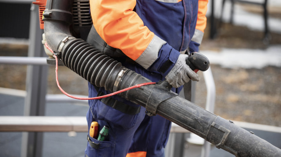 Kuvassa on henkilö, joka pitää lehtipuhallinta käsissään. Hänellä on päällään kirkkaan oranssi ja sininen työhaalari sekä hanskat. Kuvan keskipiste on lehtipuhaltimessa ja henkilön työvarusteissa. Tausta on hieman epätarkka, mutta viittaa ulkotyöympäristöön, mahdollisesti puisto- tai katualueella. Tunnelma on toiminnallinen, ja kuva korostaa työntekoa ulkona.