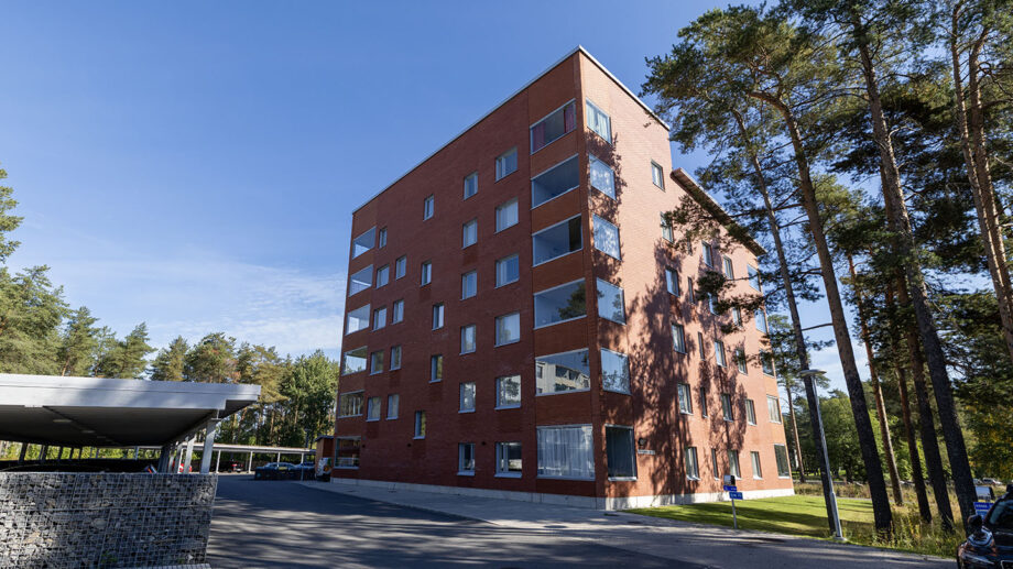 Punatiilinen kuusikerroksinen kerrostalo, aurinkoinen julkisivu ja pysäköintipaikka edessä.