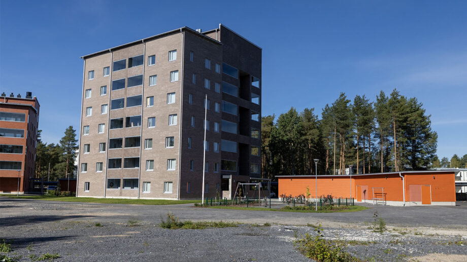 Harmaa kerrostalo ja oranssi piharakennus, sinitaivas ja avoin piha-alue.