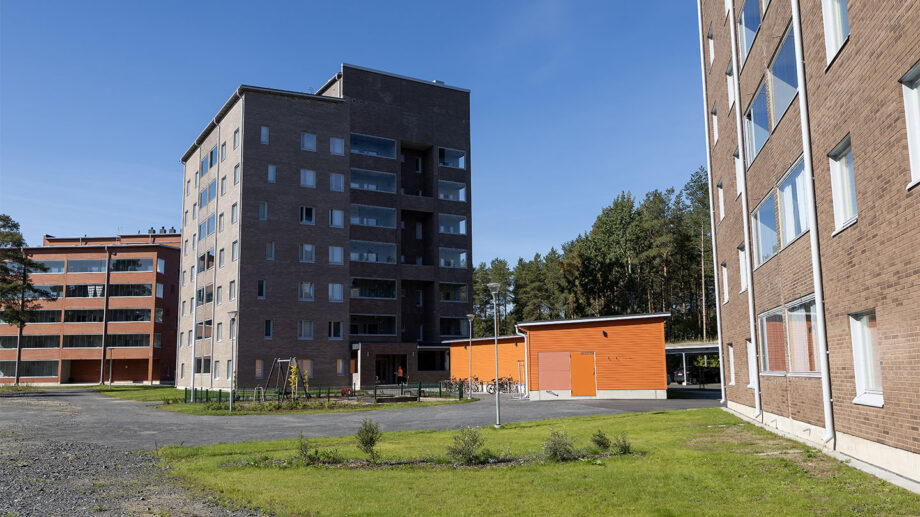 Moderni harmaa kerrostalo ja oranssi piharakennus, vihreä nurmialue edessä.