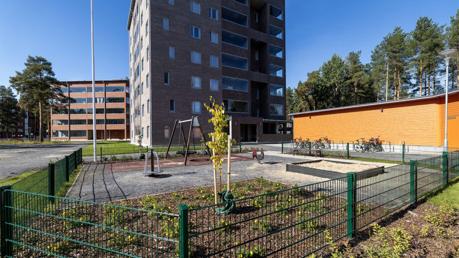 Harmaasävyinen kuusikerroksinen asuinkerrostalo, etualalla aidattu leikkipiha.