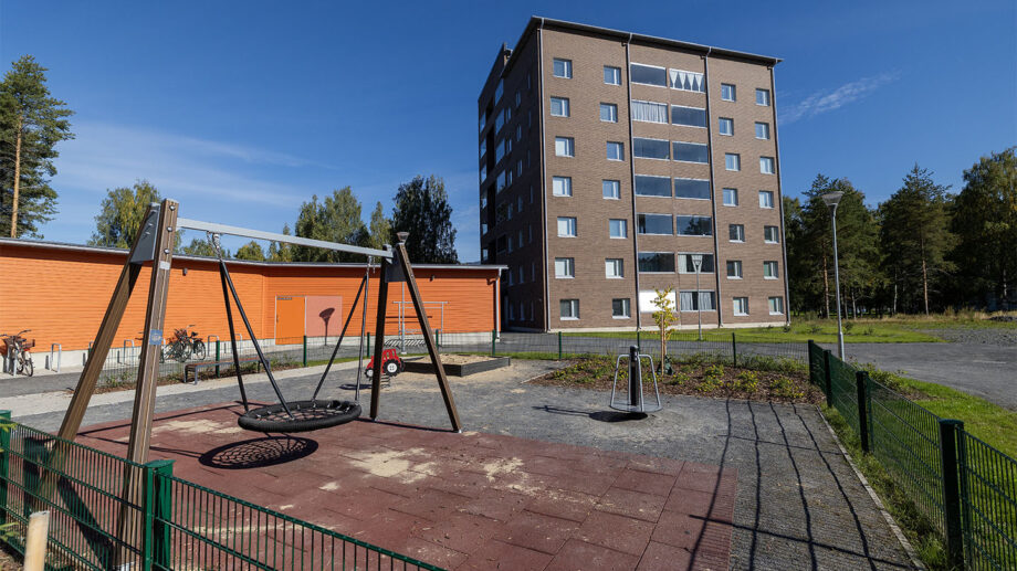 Leikkipiha ja kerrostalon julkisivu, etualalla hiekkakenttä ja keinuja.