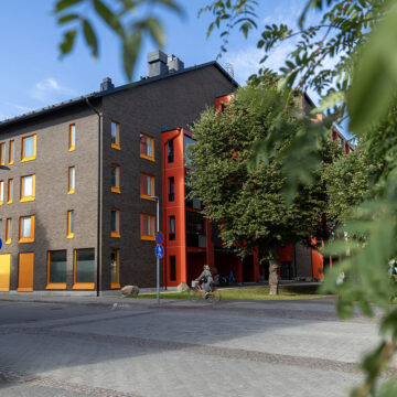 Tummasävyinen viisikerroksinen asuinkerrostalo, punaisia parvekeseiniä ja sisäänkäynti kadun varrella.