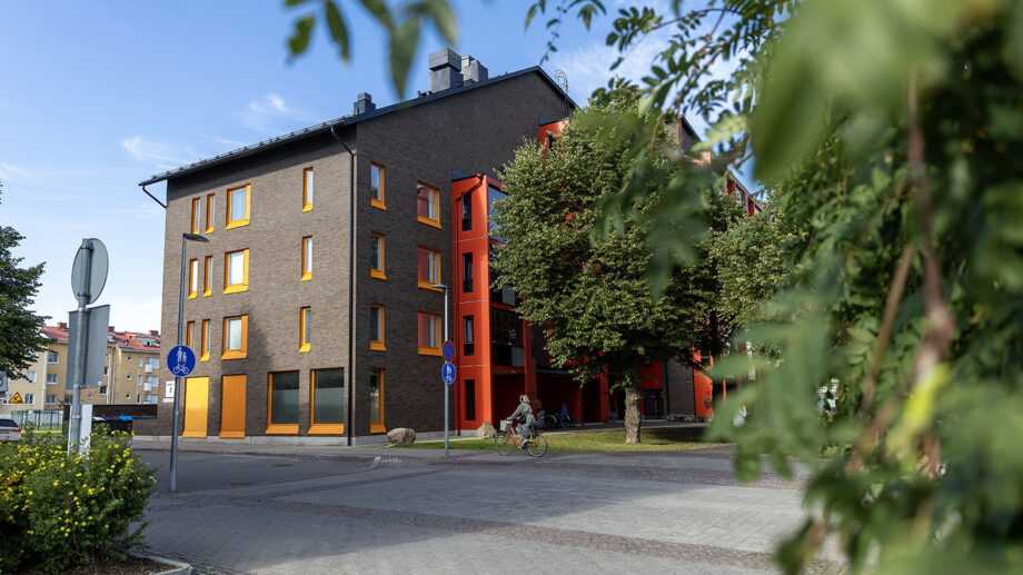 Tummasävyinen viisikerroksinen asuinkerrostalo, punaisia parvekeseiniä ja sisäänkäynti kadun varrella.