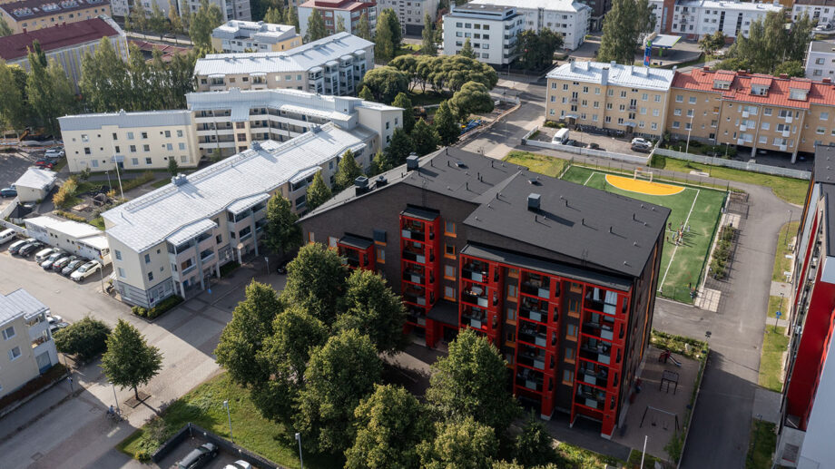 Ilmakuva kerrostalosta, jossa punaiset parvekkeet ja musta katto, ympärillä puita ja kaupunkialue.