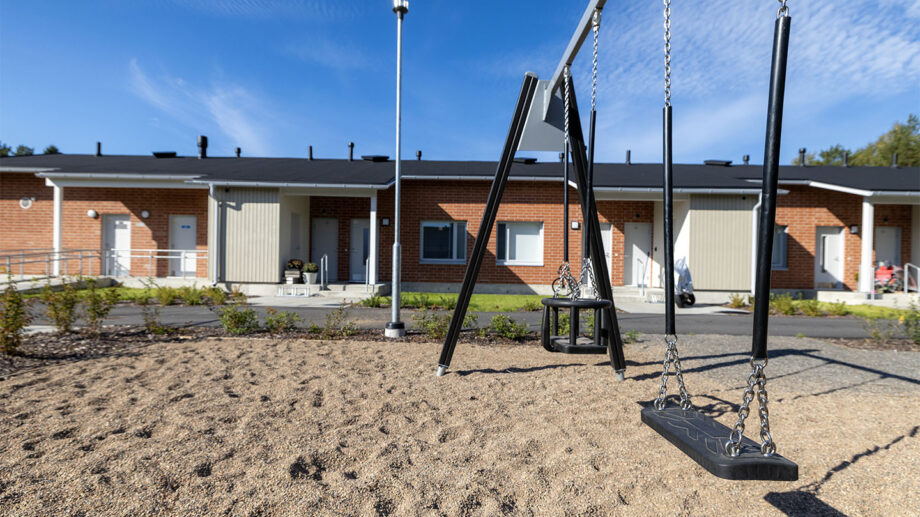 Leikkipiha rivitalon edustalla, keinu hiekkakentällä.