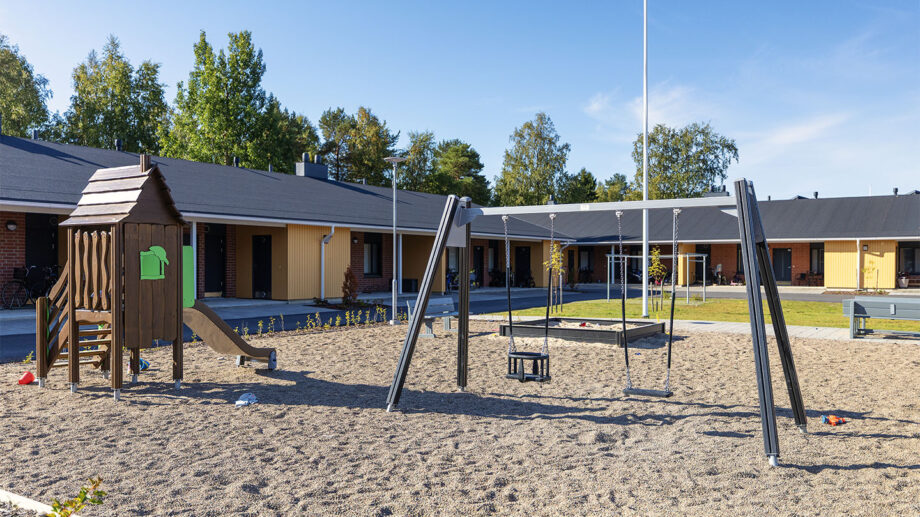 Leikkipiha ja vaaleasävyinen yksikerroksinen rivitalo, hiekka-alueella keinuja ja katos