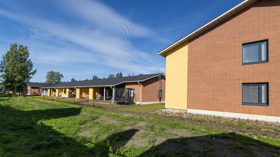 Vaaleankeltainen kaksikerroksinen rivitalo, aurinkoinen piha ja sinitaivas.