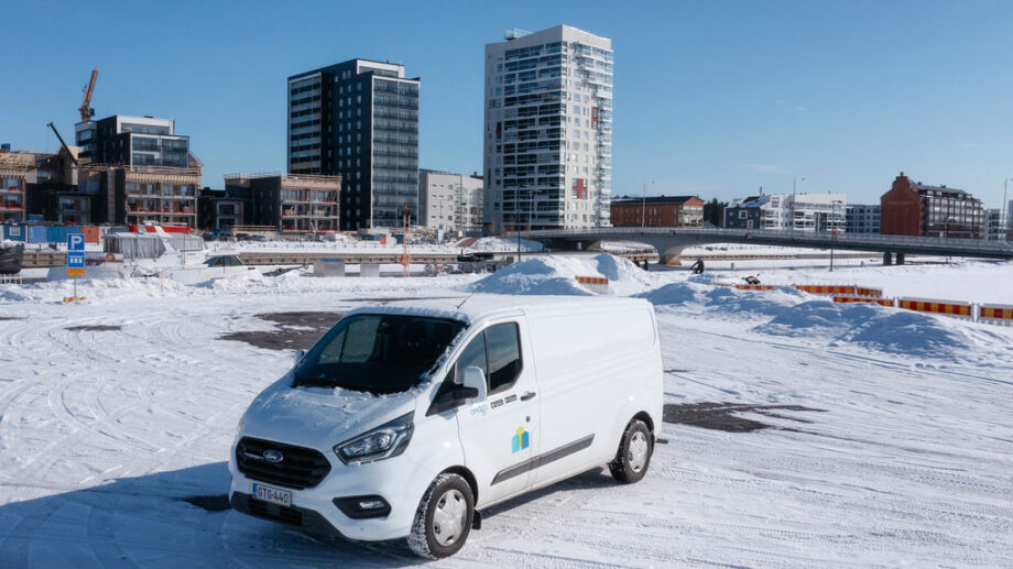 Kuvassa on valkoinen pakettiauto lumisella alueella, taustalla näkyy moderneja korkeita rakennuksia sekä keskeneräistä rakennustyömaata. Taivas on kirkkaan sininen, ja aurinko paistaa kirkkaasti, luoden selkeän talvisen maiseman. Pakettiauton kyljessä näkyy logo, ja se on pysäköity avaralle lumiselle parkkialueelle. Kuva korostaa auton käytettävyyttä sekä kaupunkimaisemaa.