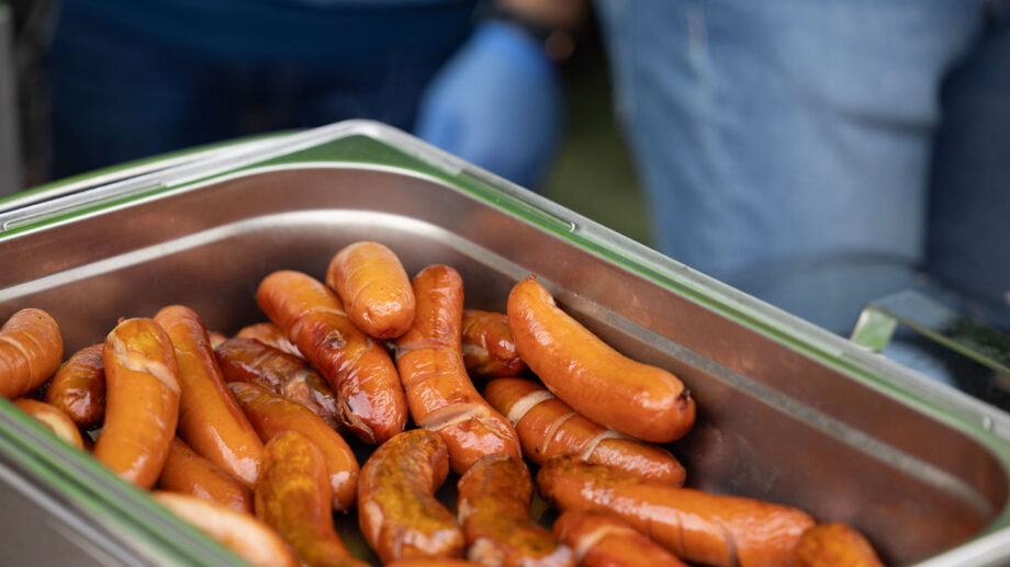 Metallisessa tarjoiluastiassa useita kymmeniä grillimakkaroita. Taustalla näkyy ihmistä ja kädessä oleva muovikäsine.