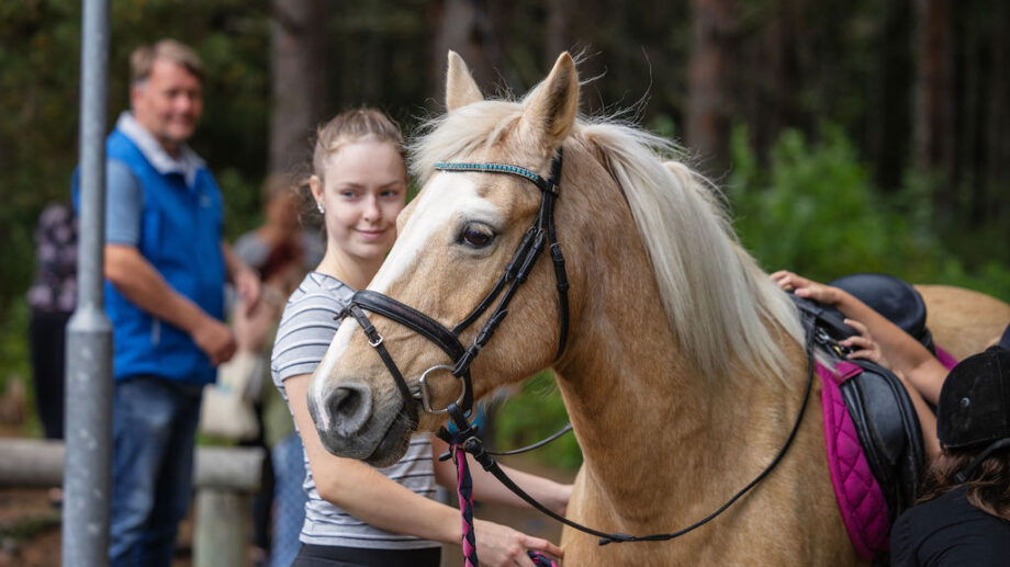 Nuori nainen pitelee vaaleaa hevosta ohjaksista hymyillen. Hevonen on satuloitu ja sen selkään on nousemassa toinen henkilö. Taustalla näkyy ihmisiä ja metsäinen ympäristö.