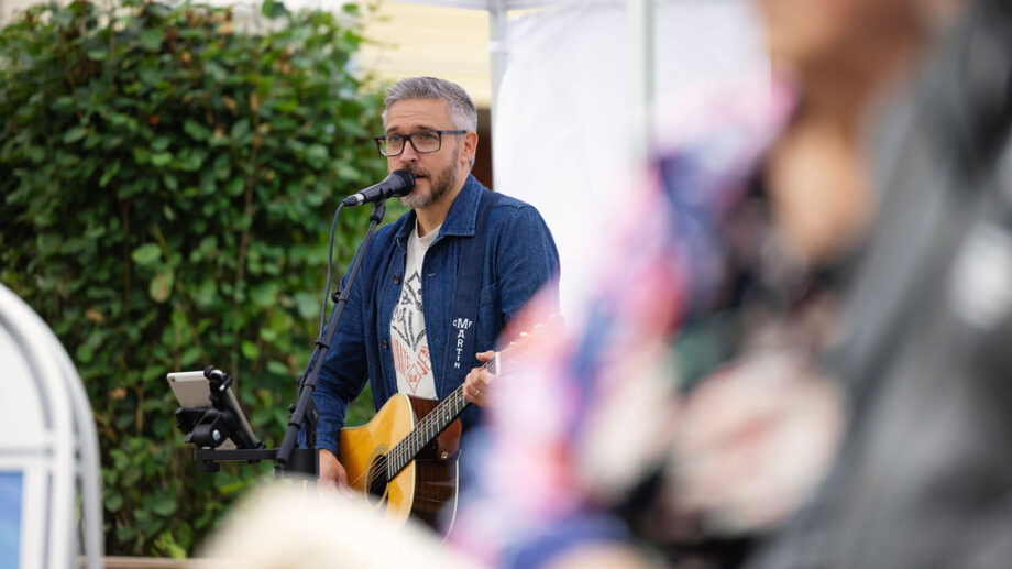 Mieshenkilö laulaa kuvassa taustalla ulkoilmatapahtumassa. Kuvan edessä häivytettynä ihimisen vartalo.