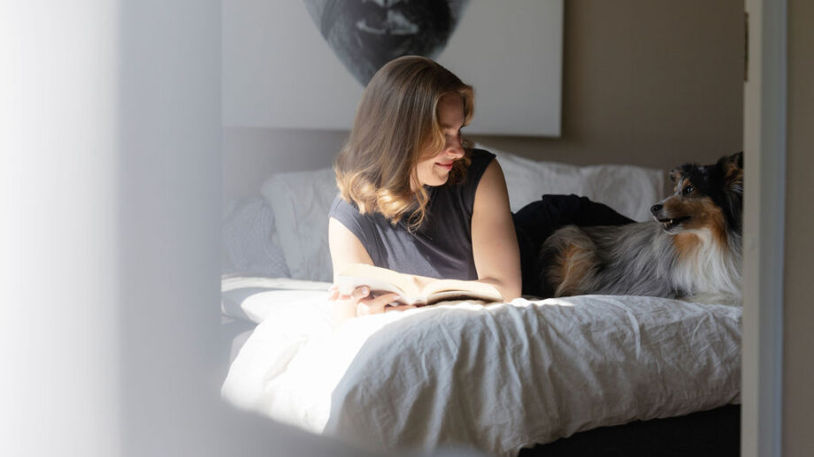 Nainen makaa sängyllä lukemassa kirjaa ja katsoo hymyillen vieressä lepäävää koiraa. Huoneeseen tulvii luonnonvaloa, ja taustalla näkyy taulu ja tyynyjä.