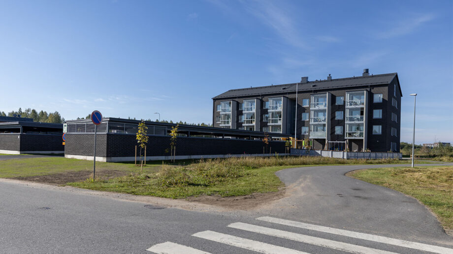 Moderni tumma asuinkerrostalo kulmasta nähtynä, etualalla suojatie ja katuympäristö.