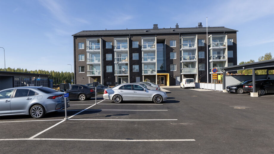Kerrostalon edustalla parkkipaikka ja kävelytie, julkisivussa lasitetut parvekkeet.