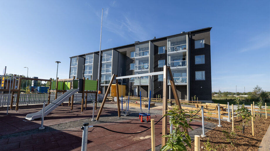 Moderni tummasävyinen kerrostalo, etualalla leikkipiha ja kiipeilytelineitä.