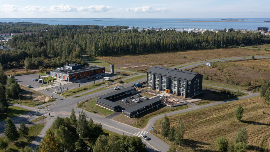 Ilmakuva mustasävyisestä nelikerroksisesta asuinkerrostalosta avoimella alueella, taustalla meri ja metsä.