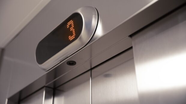 Hissin ovet ja kerrosnäyttö — Hissin yläpuolella näkyy digitaalinen näyttö, jossa lukee numero 3 oranssilla värillä, mikä ilmaisee, että hissi on kolmannessa kerroksessa.