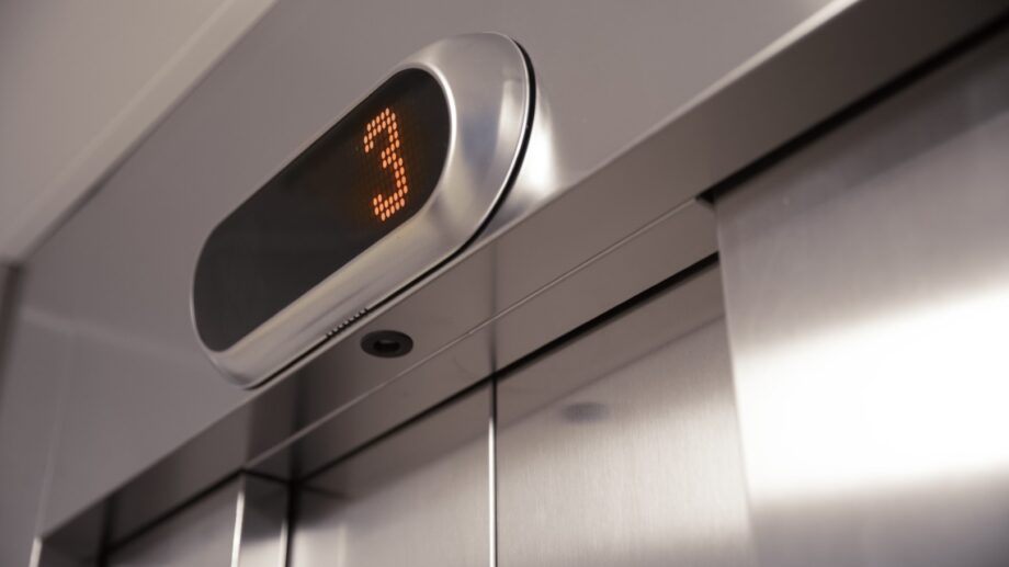 Hissin ovet ja kerrosnäyttö — Hissin yläpuolella näkyy digitaalinen näyttö, jossa lukee numero 3 oranssilla värillä, mikä ilmaisee, että hissi on kolmannessa kerroksessa.