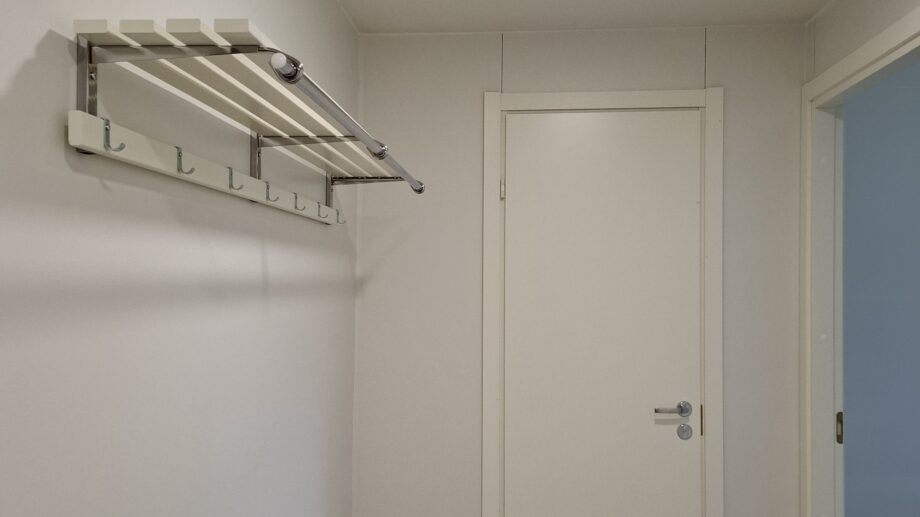 Valoisa eteinen, jossa on valkoinen ovi, seinään kiinnitetty naulakko ja hylly sekä vaalea seinä- ja kattopinta.