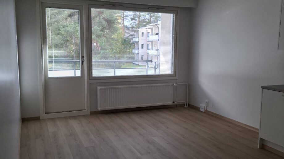 Valoisa huone, jossa on vaalea puulattia, valkoinen paneelikatto ja suuret ikkunat, jotka avautuvat parvekkeelle ja näkymään taloyhtiön piha-alueelle.