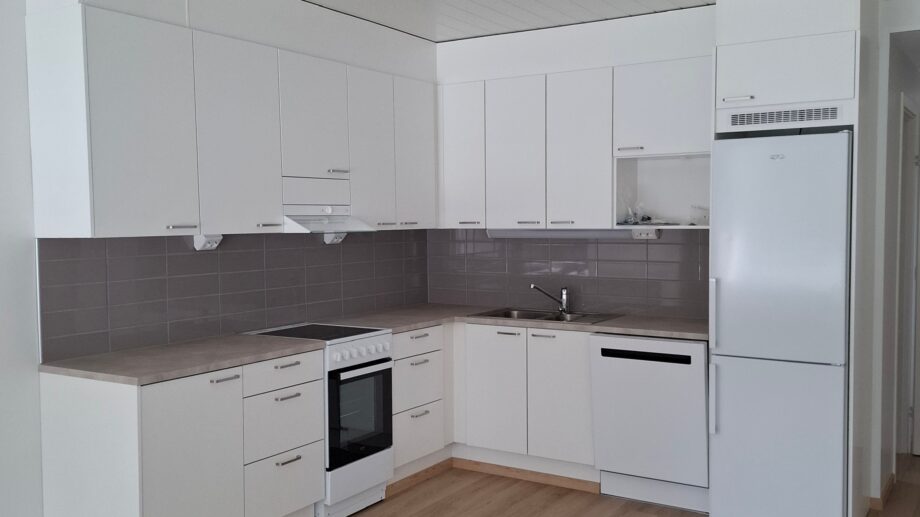 Moderni vaaleasävyinen keittiö, jossa on valkoiset kaapistot, harmaa laattavälitila, liesi, astianpesukone, jääkaappi-pakastin ja vaalea vinyylilattia.