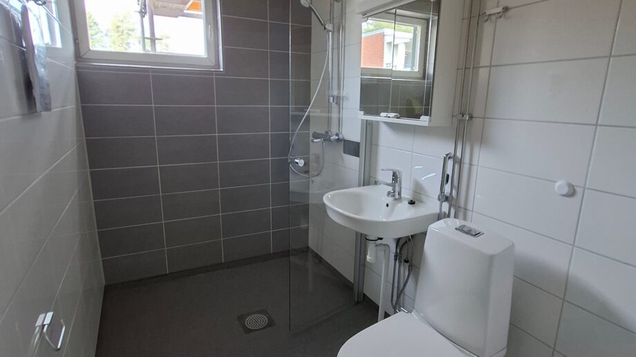 Laatoitettu kylpyhuone, jossa on wc-istuin, pesuallas peilikaapilla, suihku lasiseinällä ja pieni ikkuna, sekä puupaneelikatto.