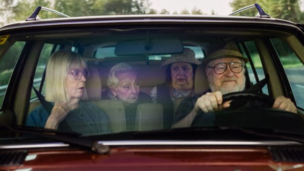 Neljän hengen seurue istuu autossa: kuljettaja ja kolme matkustajaa katsovat eteenpäin vaihtelevin ilmein, kuin yllättyneinä tai keskustellen.