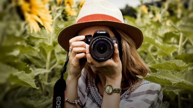 kuvassa lierihattupäinen nainen, jolla järjestelmäkamera kasvojen edessä. Taustalla vihreä pelto.