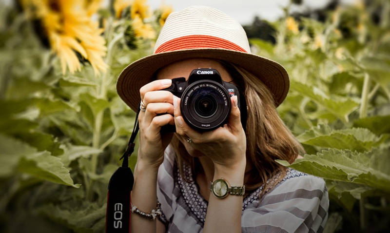 kuvassa lierihattupäinen nainen, jolla järjestelmäkamera kasvojen edessä. Taustalla vihreä pelto.