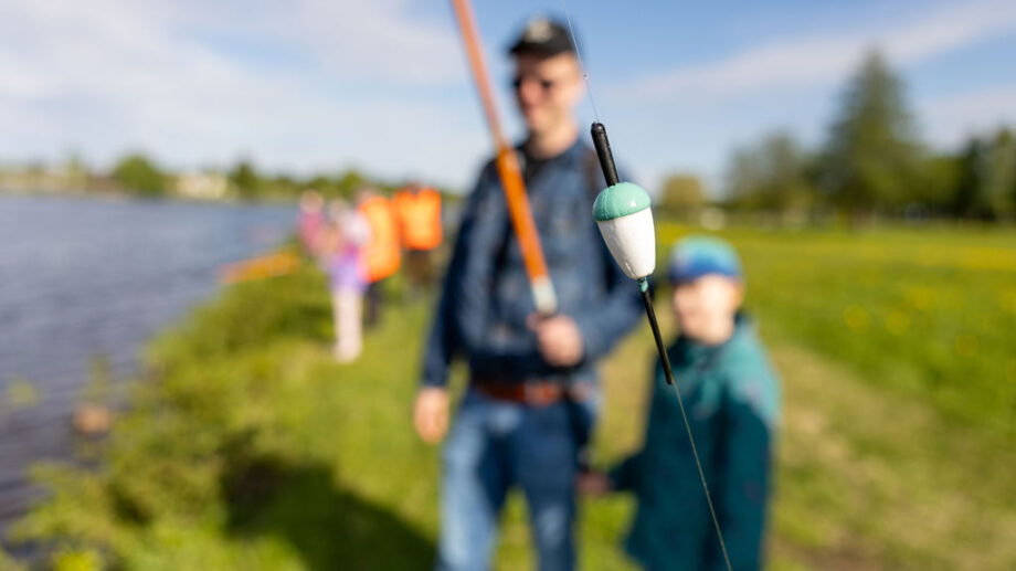 Edustalla ongen koho tarkennettuna. Taustalla mies ja poika, jotka nurmialueella vesistön vieressä. Taustalla myös muita onkijoita.